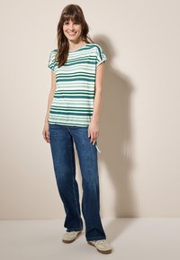 Camiseta de rayas verdes y blancas con mangas cortas, combinada con vaqueros azules holgados y zapatos de color claro. Detalle notable de lazo en el costado de la camiseta.