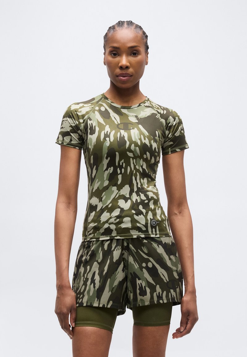 Γυναίκα που στέκεται και κοιτάζει μπροστά, φοράει κοντομάνικο πουκάμισο και σορτς με πράσινο και καφέ αφηρημένο καμουφλάζ, πάνω από ελαστικά εσώρουχα σε ελιάς χρώμα.