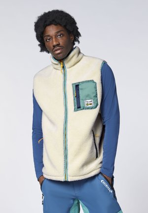 Man with curly hair wearing a cream fleece vest with green pocket over a blue long-sleeve shirt and blue pants with hands in pockets.