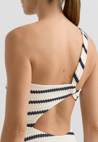 Merevítő nélküli egyrészes fürdőruha texturált anyagból, krémszínű és tengerészkék csíkokkal, nyitott hátú kivitelben, és csavart vállpánt részlettel.