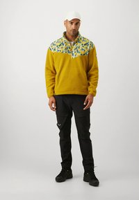 Κίτρινο φλις πουλόβερ με γεωμετρικό γιακά σε μπλε και κίτρινες αποχρώσεις, συνδυασμένο με μαύρο παντελόνι cargo και μαύρα παπούτσια.