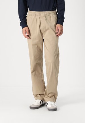 Mann trägt beigefarbene Hosen mit elastischem Bund, weiße und graue Turnschuhe und ein dunkles langärmeliges Hemd, steht vor einem einfarbigen Hintergrund.