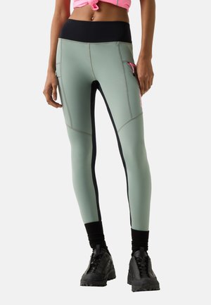 Femme portant des leggings verts et noirs avec des poches latérales, des chaussettes noires, des baskets noires et un crop top rose noué, debout sur fond blanc.