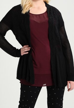 Femme portant un haut bordeaux transparent superposé avec un cardigan en maille noir et un pantalon noir à petits pois blancs, main sur la hanche.