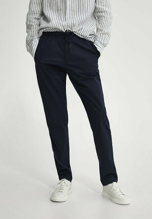 Personne en pantalon noir à cordon, baskets blanches et chemise blanche à rayures verticales noires, debout les mains dans les poches.