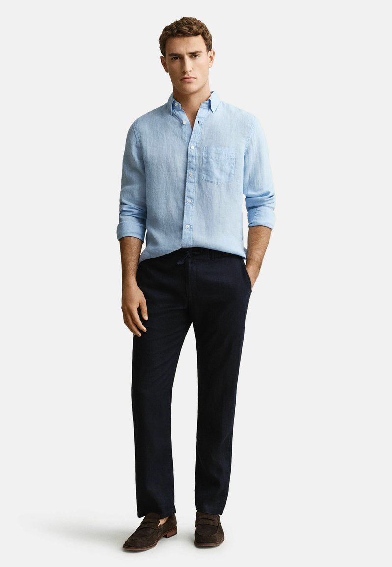 Junger Mann stehend, trägt ein hellblaues Hemd mit langen Ärmeln, dunkle Hose und braune Loafers, mit einer Hand in der Tasche.