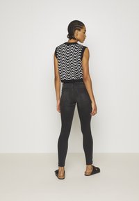 Top preto sem mangas com um padrão branco ondulado nas costas, combinado com jeans pretos justos e sandálias pretas de deslizar. Textura de tecido suave.