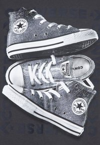 Sivi bleščičasti Converse superge z visokim zgornjim delom, belimi gumijastimi nosilci, črnimi črtami in okroglo blagovno znamko na strani.