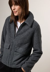 Veste en polaire grise avec un extérieur texturé, fermeture éclair à l'avant et poches poitrine doubles avec boutons-pression, complétée par un col.