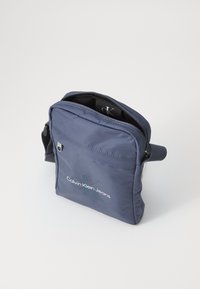 Sötétkék Calvin Klein Jeans keresztpántos táska cipzáras főrekesszel és elülső zsebbel, sima fehér háttér előtt.