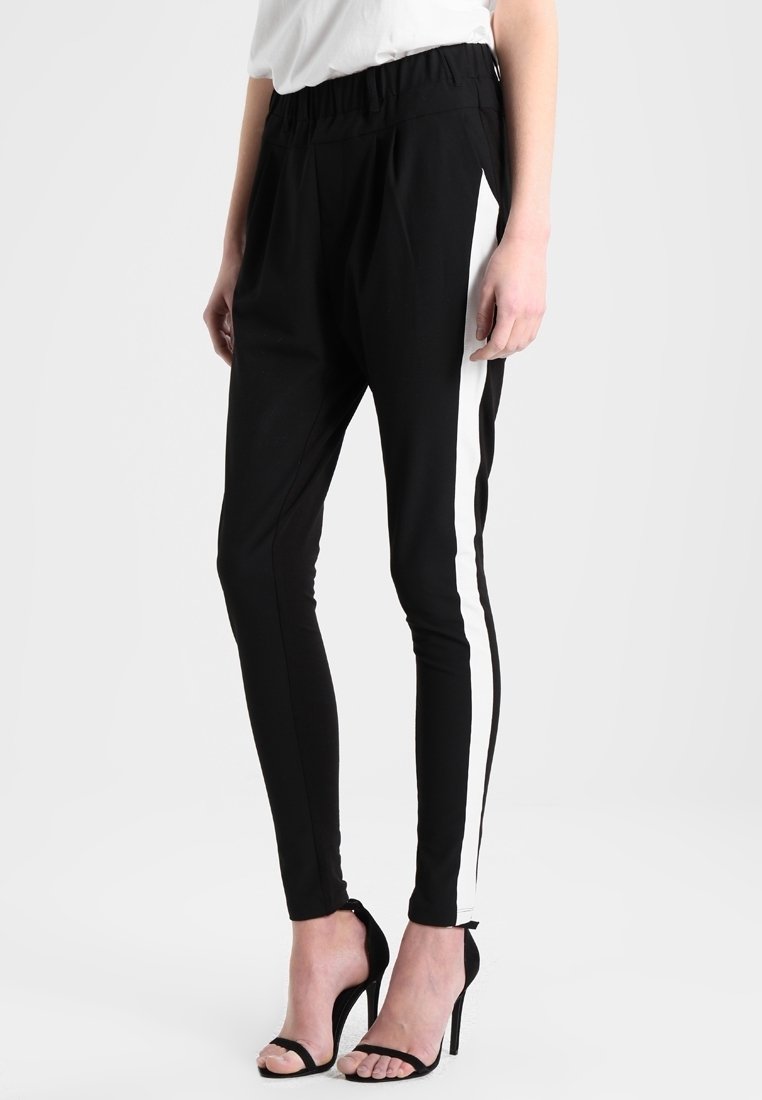 Personne portant un pantalon noir slim avec des bandes blanches sur les côtés et des sandales à talons noires, debout devant un fond uni.