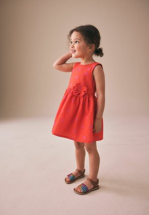 Junges Mädchen lächelt und schaut nach rechts, trägt ein ärmelloses strukturiertes rotes Kleid mit Blumendetail und bunte Sandalen, steht auf einem beigen Boden.