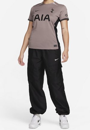 Maillot de sport gris avec le logo noir "AIA" et l'emblème de Tottenham, associé à un pantalon noir ample et des baskets blanches Nike, arborant un design épuré.