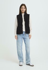 Zwarte fleece vest met een rits aan de voorkant, twee zijzakken, gedragen over een lichtgekleurde trui, gecombineerd met lichtblauwe jeans en sneakers.