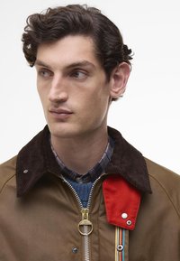 Brown utility jacket with a corduroy collar, red accent, and multicolored stripe detail. Features a zipper closure and sturdy fabric texture.