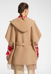 Poncho beige intrecciato con cappuccio, legato in vita, con dettagli a frange all'orlo. Il modello indossa una camicia rossa con motivo sottostante.
