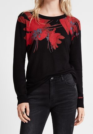 Femme portant un pull noir avec des motifs floraux rouges sur la poitrine et les épaules, associé à un jean noir et des bagues aux doigts.