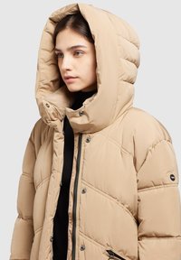 Veste doudoune beige avec une grande capuche matelassée, des coutures diagonales et des poches zippées. Comprend des boutons-pression et une texture lisse.
