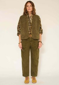 Pantalons cargo vert olive avec poches latérales, un gilet marron texturé et une chemise à carreaux à boutons. Des chaussures à lacets marron complètent le look.