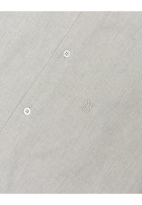 Jasnoszara koszula zapinana na guziki z teksturowanej tkaniny, z białymi plastikowymi guzikami i małym haftowanym logo po lewej stronie.