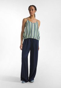 Kobieta stojąca, ubrana w zielono-niebieską pionowo paskowaną, bezrękawnik, luźne granatowe spodnie i jasnoniebieskie buty na białym tle.