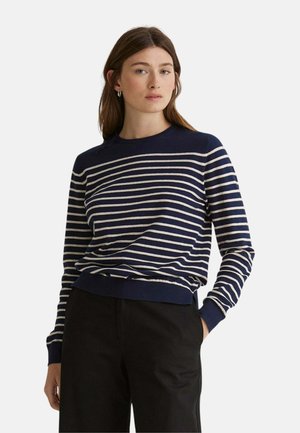 Mujer con el pelo largo y castaño, vestida con un suéter a rayas azul marino y blanco y pantalones negros, de pie con una mano en el bolsillo sobre un fondo gris.
