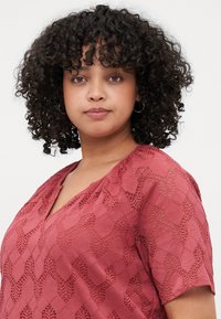 Blusa rosa con scollatura a V, maniche corte e un motivo in pizzo intricati che presenta forme geometriche e ritagli.