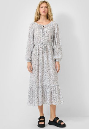 Kobieta ubrana w białą, kwiecistą sukienkę midi z długim rękawem, ze sznurkiem w talii i falbaną na dole, do tego czarne sandały na platformie.