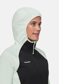 Kaptrui met een twee-kleurig ontwerp in licht muntgroen en zwart, met een gestructureerde stof en een voorkant met rits die bevestigt is met een trekkoord.