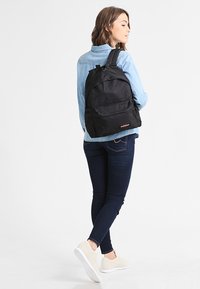 Mochila de nylon negra con correas acolchadas y un bolsillo frontal, que presenta un parche de logo. La bolsa tiene una forma redondeada y una textura suave.
