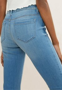 Jeans de mezclilla azul con una textura suave, cintura alta y dos bolsillos traseros. Costuras en un color contrastante a lo largo de las costuras.