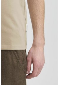 Beige pamut póló oldalsó címkével, barna vászonnadrággal párosítva. Kiemelt figyelmet kap a sima anyag és a laza szabás részletei.