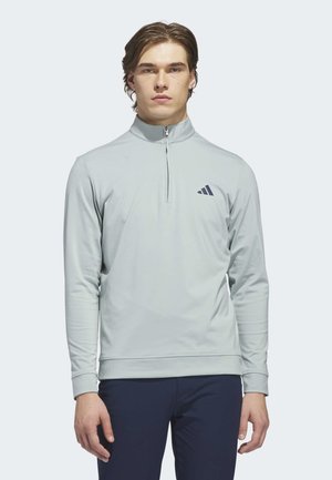 Jeune homme portant un pull léger gris clair à fermeture éclair courte avec un logo sur le côté gauche de la poitrine et un pantalon foncé, debout devant un fond uni clair.