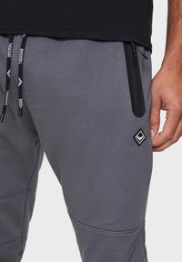 Graue Sweatpants mit schlanker Passform, ausgestattet mit einer Reißverschluss-Tasche, schwarzen Kordeln und einem rautenförmigen Logo-Patch am Oberschenkel.