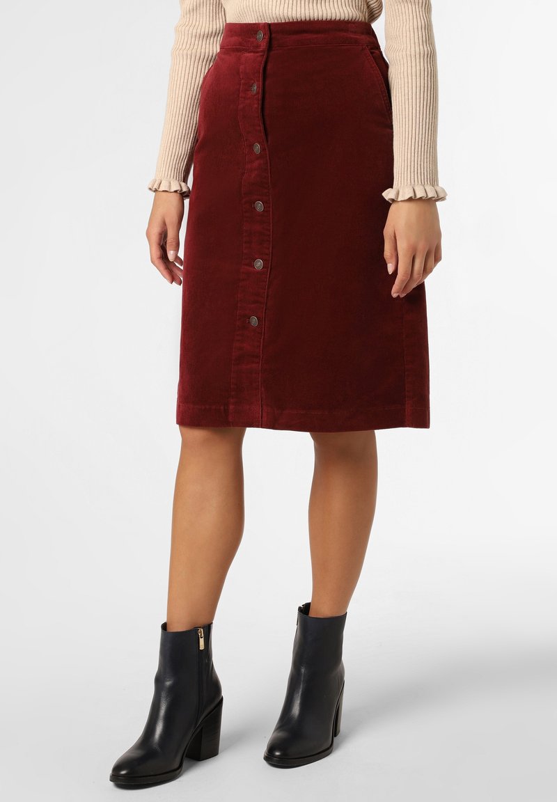 Femme portant une jupe bordeaux mi-longue à boutons sur le devant, un pull côtelé beige et des bottines à talons noires, sur un fond uni.