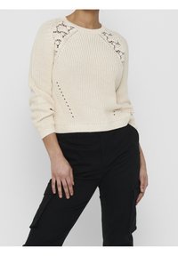 Krämfärgad stickad tröja med ett texturerat mönster, raglanärmar och en åtsittande fåll. Har öppet stickdekor på axlarna.