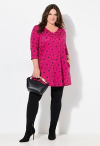 Rosa floral tunika med sorte tusindfryd mønstre, trekvart ærmer, parret med sorte leggings og ankelstøvler, holder en sort håndtaske.