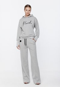 Jasnoszara bluza z kapturem z czarnym logo w formie napisu, w połączeniu z dopasowanymi, luźnymi spodniami dresowymi z sznurkiem i bocznymi kieszeniami. Gładka tekstura.