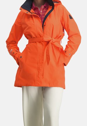 Kobieta ubrana w jasnopomarańczowy płaszcz przeciwdeszczowy z kapturem i paskiem, nałożony na różową koszulę z kołnierzykiem i białe spodnie.