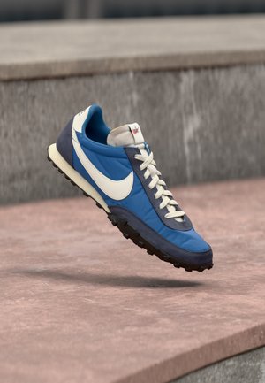 Baskets Nike bleues et marine avec le logo swoosh blanc et des lacets blancs, flottant au-dessus d'une surface en pierre avec un mur en pierre grise en arrière-plan.
