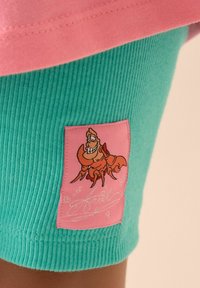 Teal ribbede shorts har en pink lap med en broderet tegneserielobster. Materialet er blødt og let tekstureret.