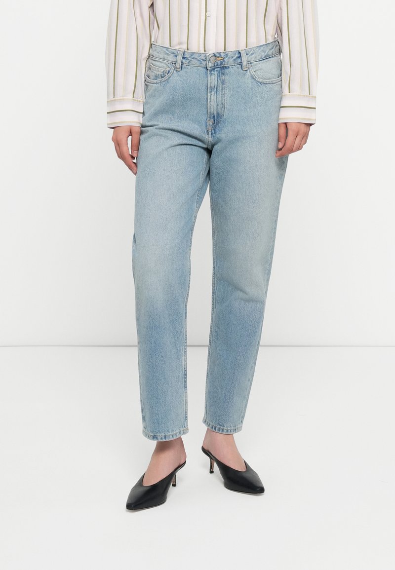 Blugi din denim bleu deschis cu croi drept, având buzunare frontale, închidere cu nasture și ușoară decolorare, asortați cu pantofi cu toc înalt negri.