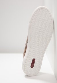 Semelle d'une chaussure en caoutchouc blanc avec un motif de bande de roulement triangulaire et un logo Rieker rouge, inclinée contre un mur blanc sur une surface plate.