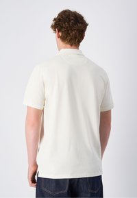 Cremefarbendes Poloshirt mit kurzen Ärmeln und Kragen, in einfarbigem Design und geradem Schnitt, kombiniert mit dunklen Jeans.