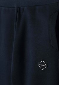 Detailný záber na tmavomodré tepláky s bočnou vreckom a malou nášivkou vo forme diamantu značky Hackett.