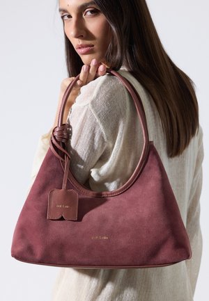 Femme aux longs cheveux bruns portant un haut en maille blanche tient un sac à main en daim rose foncé sur son épaule devant un fond uni.