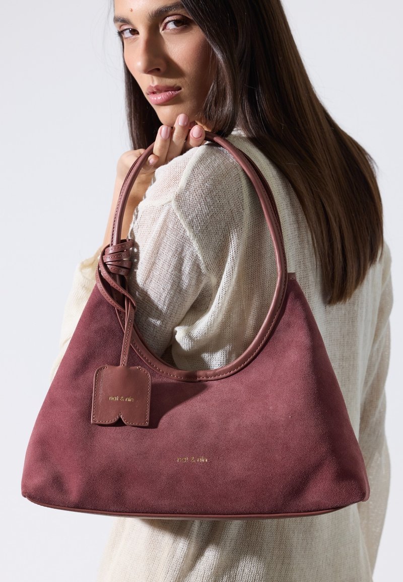 Femme aux longs cheveux bruns portant un haut en maille blanche tient un sac à main en daim rose foncé sur son épaule devant un fond uni.