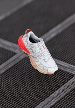 Baskets de course blanches avec des accents rouges, tige en maille texturée, semelle rembourrée et logo de marque prominent sur le côté. Conception légère, tread visible.