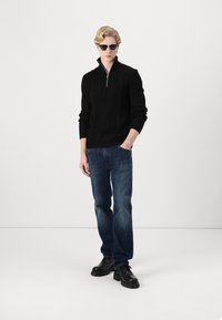 Pull en maille noire avec col zippé, associé à un jean bleu foncé et des chaussures noires à lacets. Les accessoires comprennent des lunettes de soleil sombres.