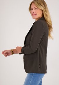 Donkergroene op maat gemaakte blazer met lange mouwen, knoopsluiting en zijsplitten, gecombineerd met lichte denim jeans. Soepele stofstructuur.
